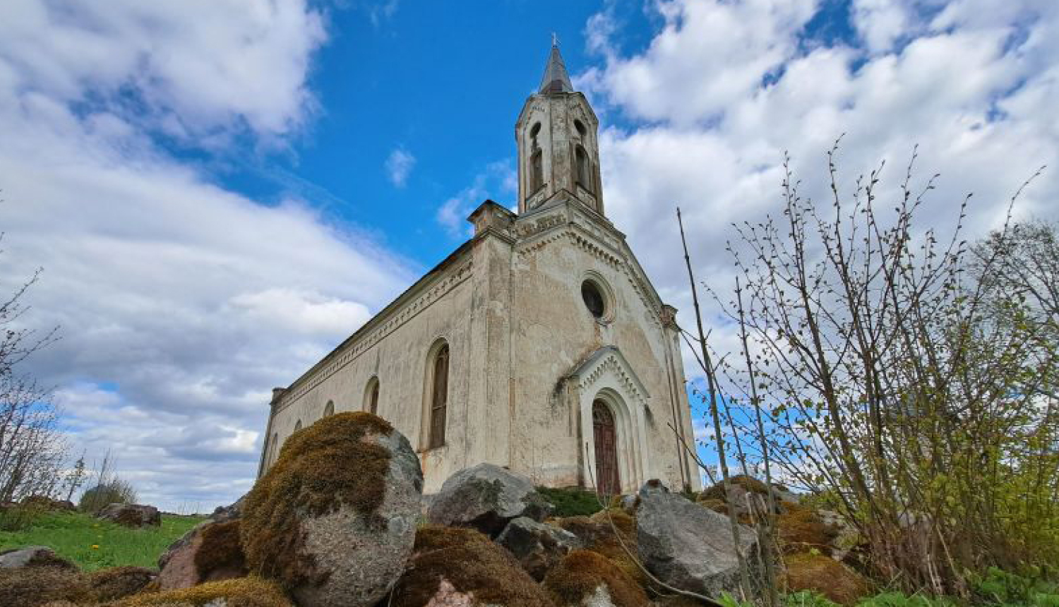 Pelēcīgi baltā Kaldabruņas baznīca pavasarī, fonā mākoņainas debesis, priekšplānā akmeņu krāvumi
