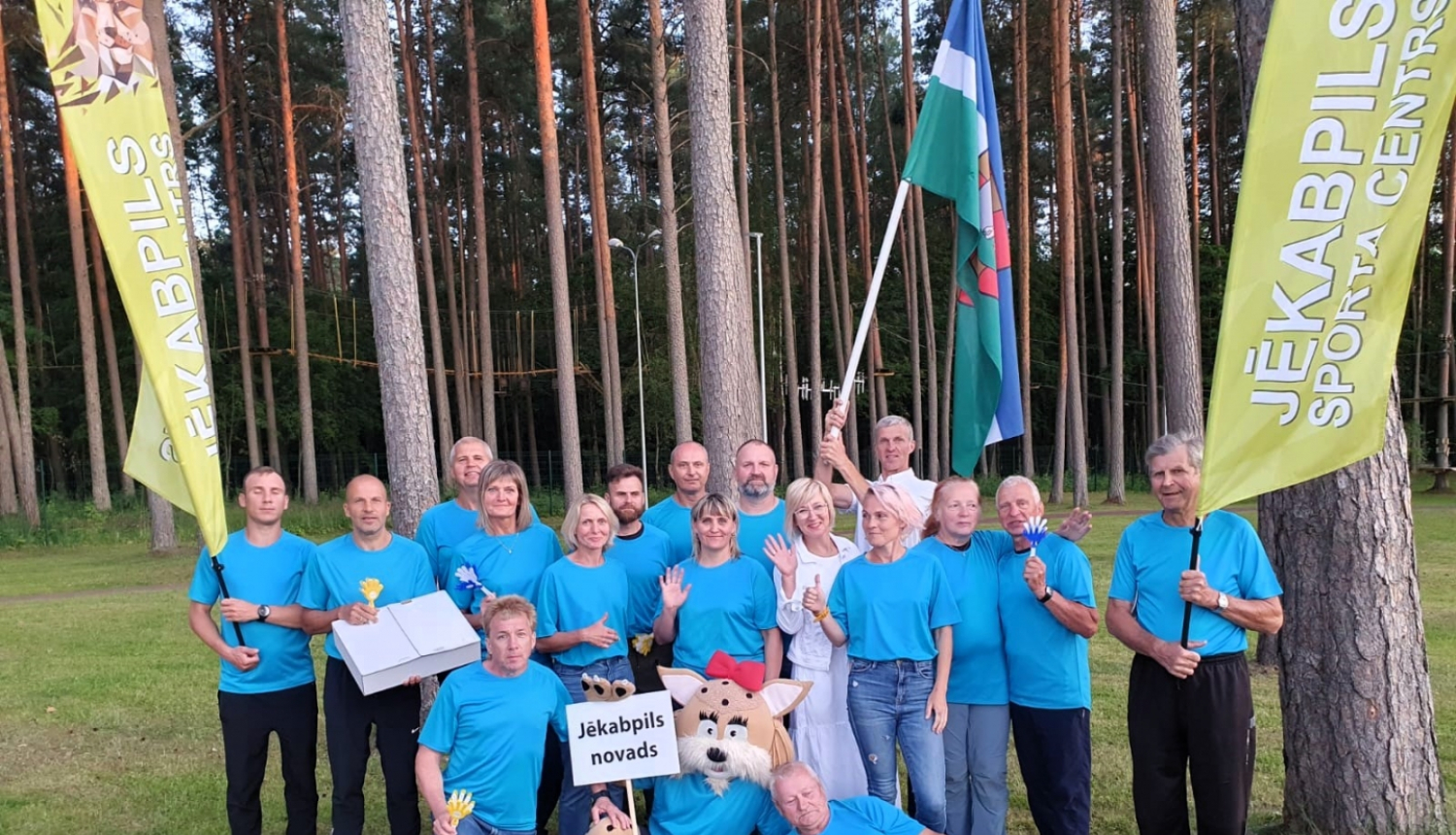 Jēkabpils novada vieglatlētu, kas piedalījās sacensībās, kopbilde ar ziliem krekliņiem, fonā novada karogs