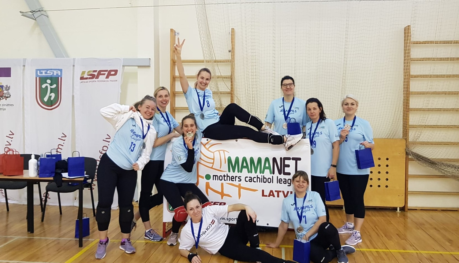 Mamanet komanda sporta zālē pozē kopbildei, turot pa vidu rokās mamanet plakātu