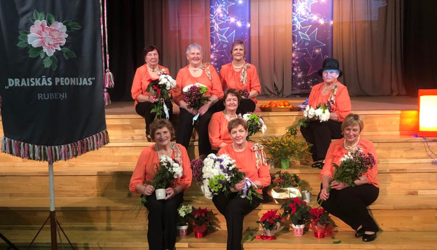 Dāmu deju kopas Draiskās Peonijas dalībnieces sēž uz Rubenes kultūras nama skatuves kāpnēm