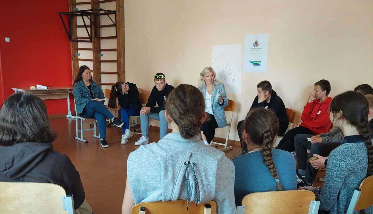 Projekta publicitātes foto - persona runā ar jauniešiem sēžot aplī krēslos