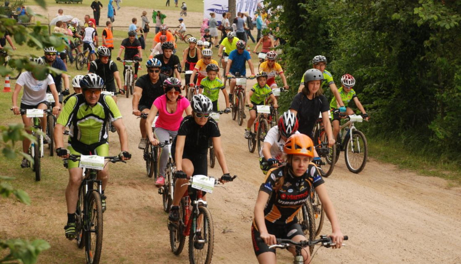 Liels daudzums velosipēdistu dažādos vecumos brauc pa zemes ceļu pretī fotogrāfam