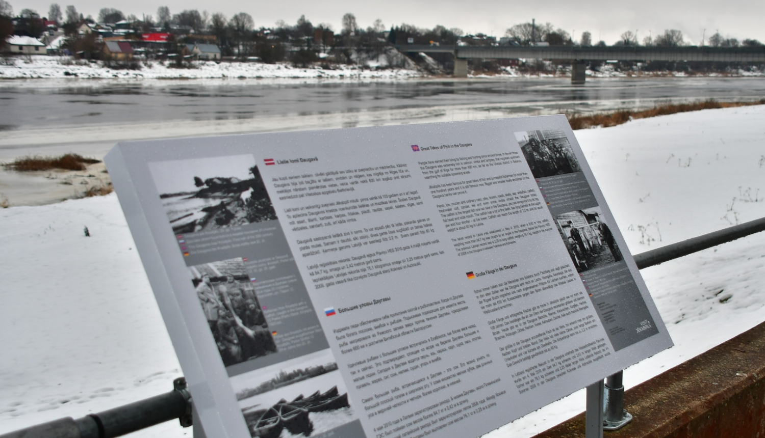 Ziemas skatā priekšplānā pelēks informatīvais stends ar tekstu dažādās valodās, fonā Daugava