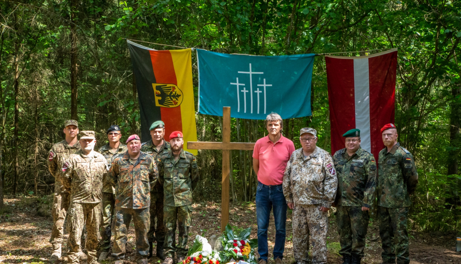 Bundesvēra karavīri, Zemessardzes un pašvaldības pārstāvis U.Skreivers stāv pie piemiņas krusta mežā, aizmugurē Vācijas un Latvijas karogi