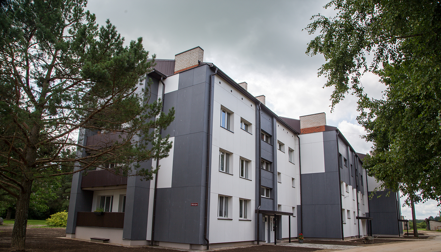 Trīs stāvu daudzdzīvokļu ēka ar jaunu pelēcīgu un baltu krāsu fasādi, lejā uzrušināta zeme, apkārt priedes