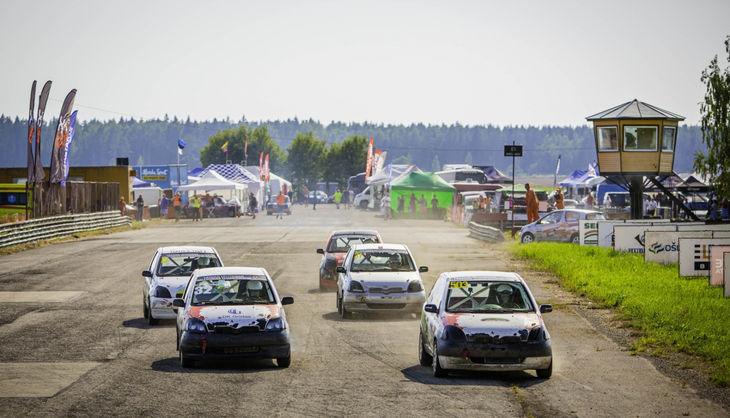 Rallijkrosa automašīnas dodas trasē, fonā tiesnešu būdiņa, teltis un skatītāji