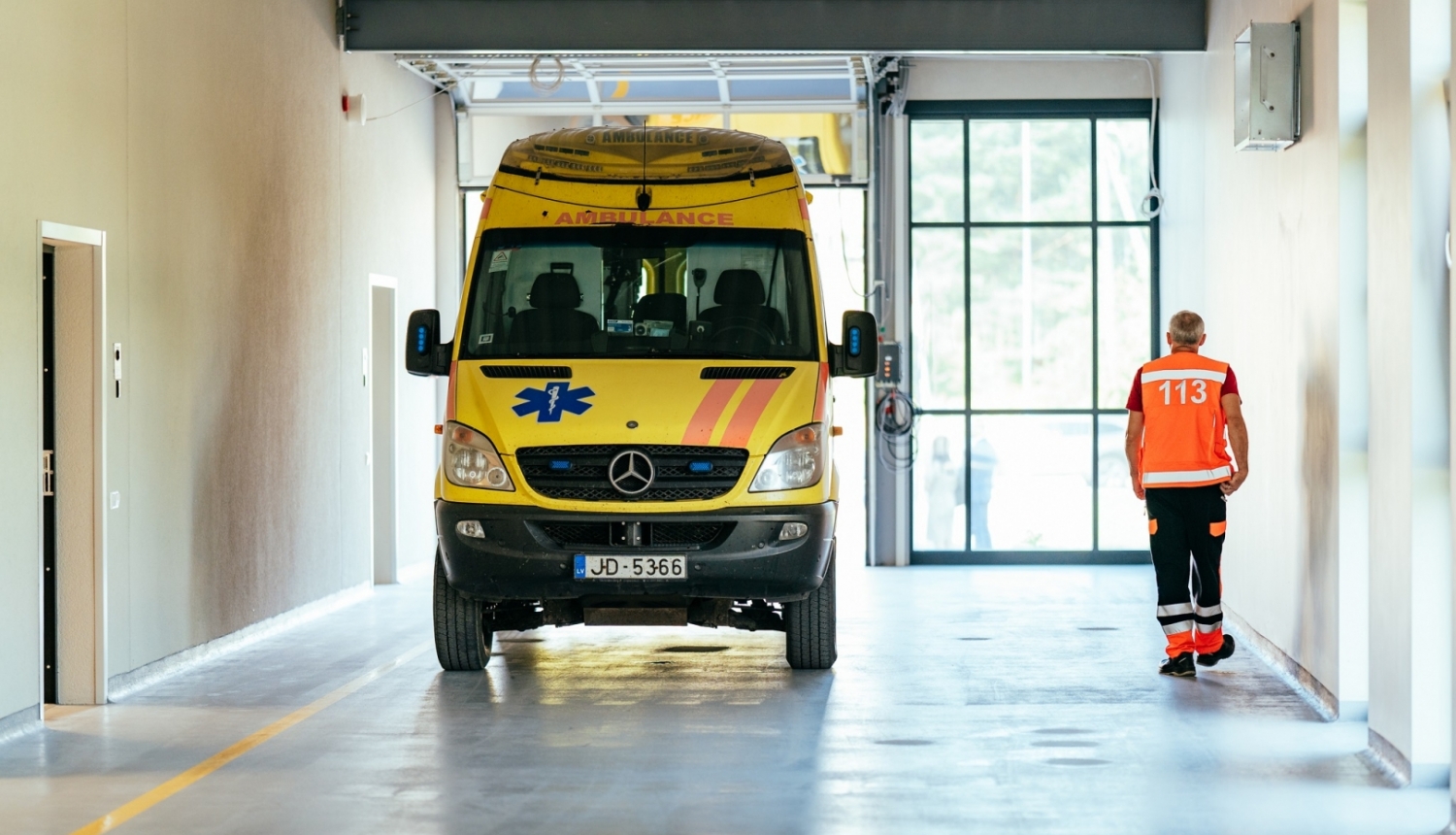 Dzeltena ātrās palīdzības mašīna telpās, blakus pa labi iet ātrās palīdzības darbinieks oranžā vestē
