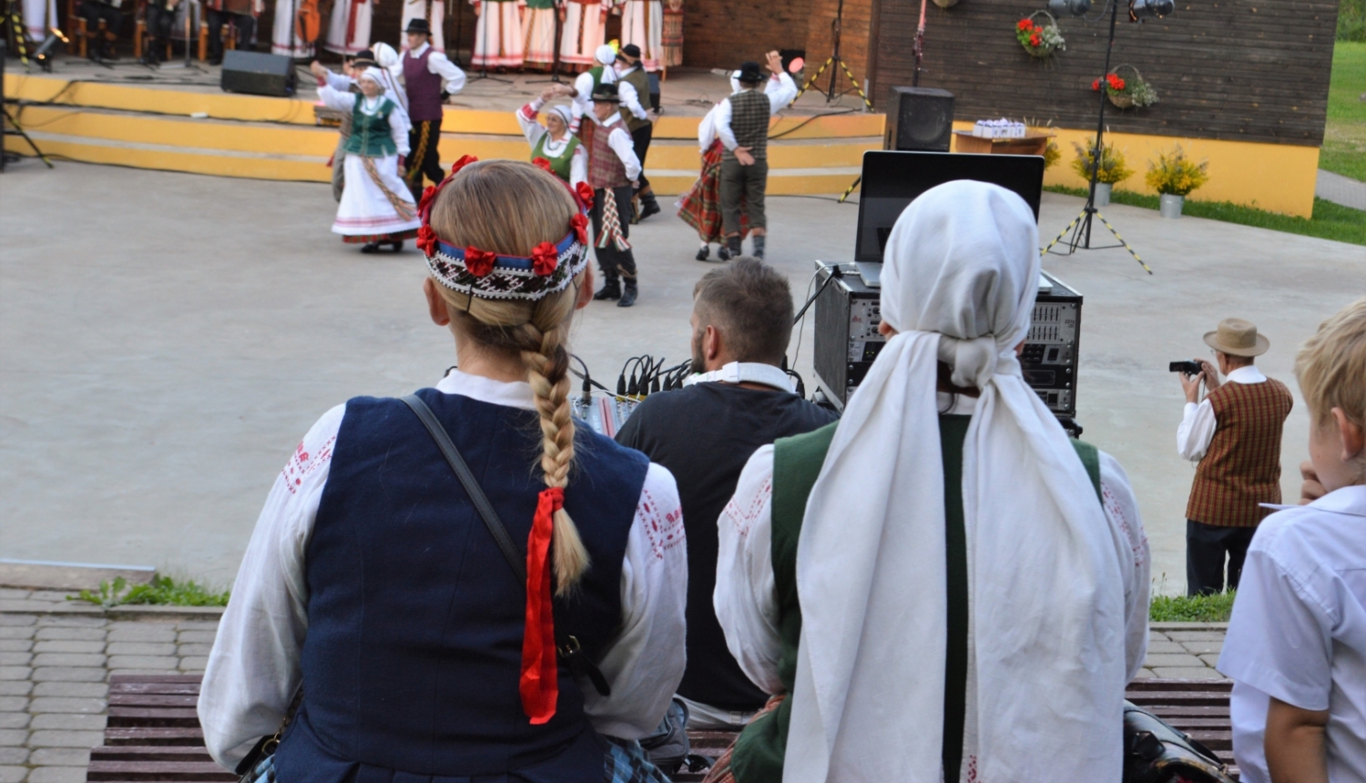Tautumeitas no mugurpuses sēž, vēro koncertu Sēlija rotā, tālumā skatuve un dejotāji