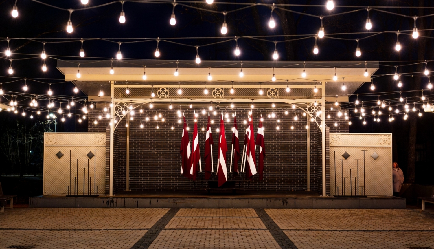 Kena parka estrāde naktī - izgaismota ar lampiņām un fonā Latvijas karogi