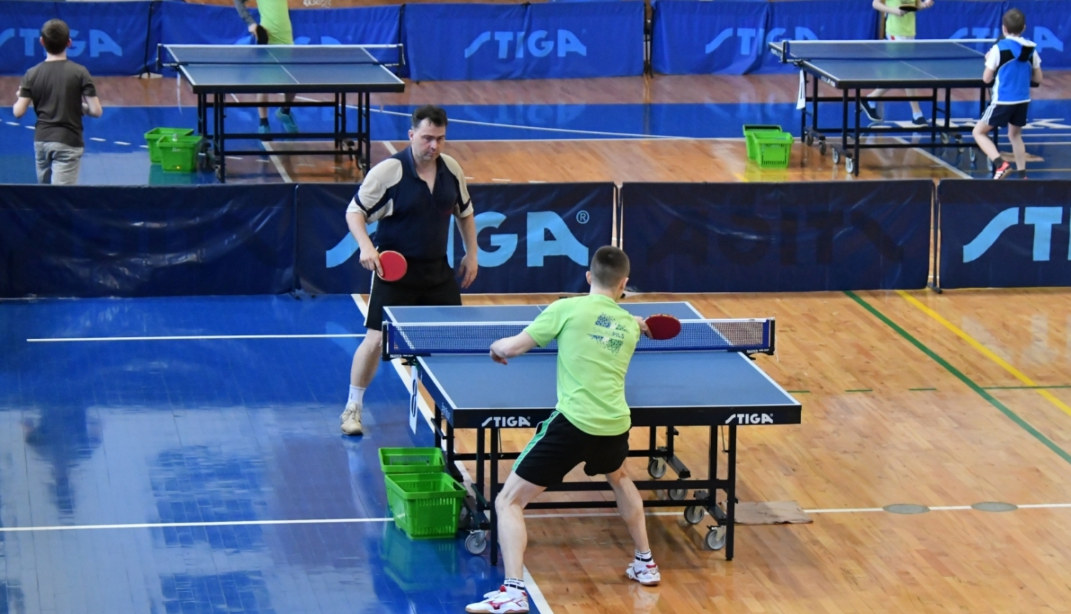 Divi vīrieši sporta zālē spēlē galda tenisu, fonā citi spēlētāji un galdi