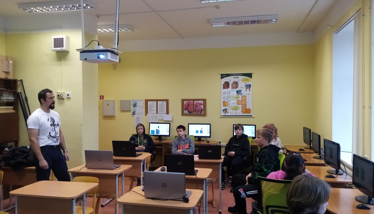 A.Bite (no kreisās) stāv un māca skolēnus, kas sēž klasē pie datoriem