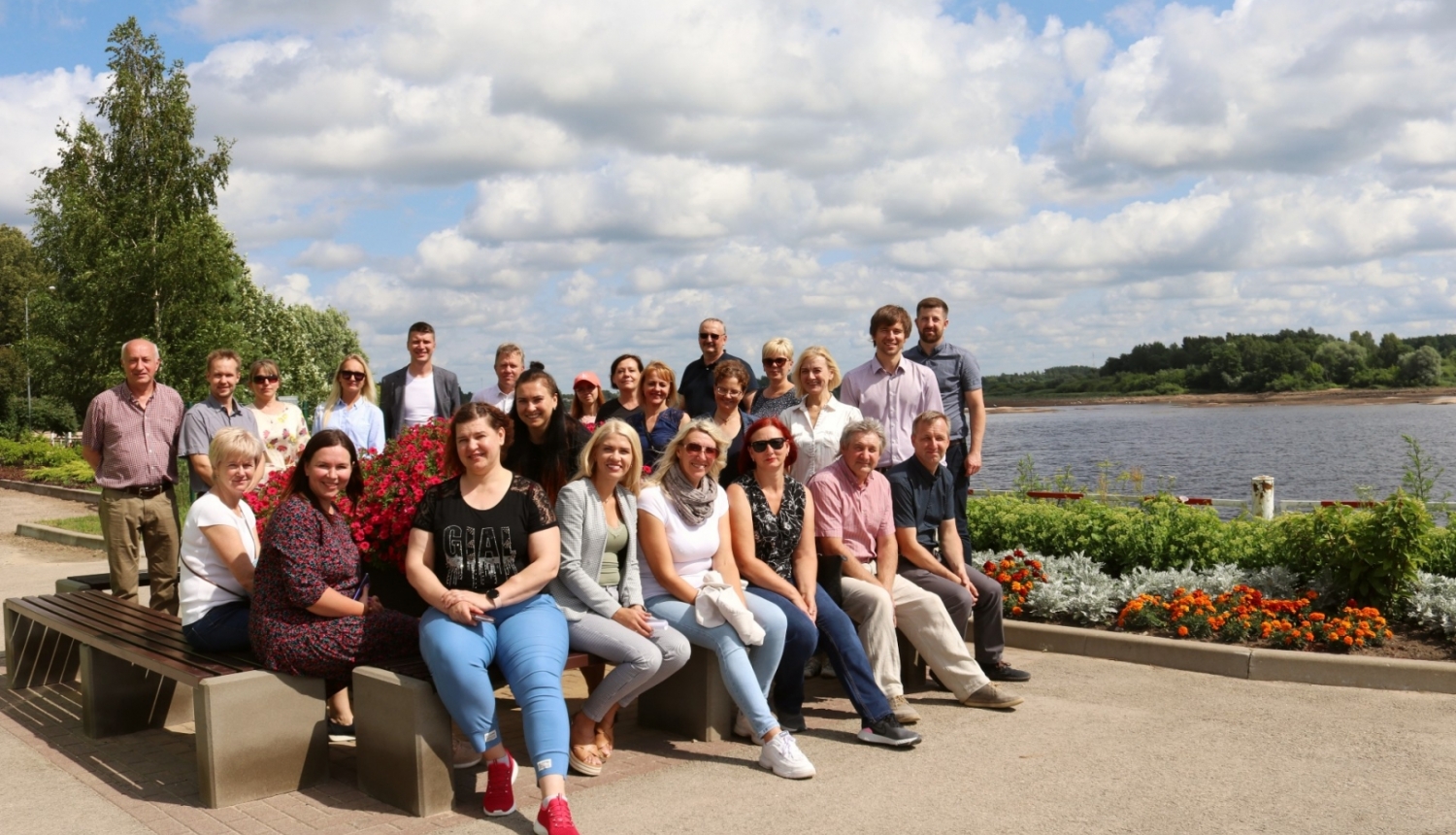 Pašvaldību dalībnieki grupā sēž uz soliņiem, fonā Daugava un puķu dobes