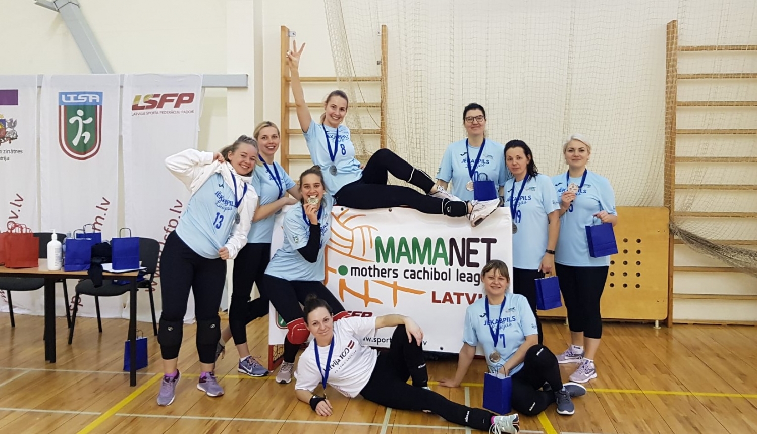 Mamanet komanda sporta zālē pozē kopbildei, turot pa vidu rokās mamanet plakātu