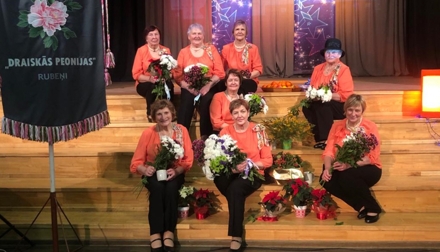 Dāmu deju kopas Draiskās Peonijas dalībnieces sēž uz Rubenes kultūras nama skatuves kāpnēm