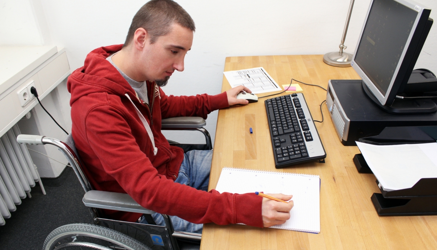 Persona ratiņkrēslā pie darba galda raksta