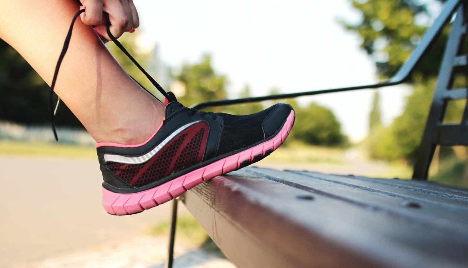 Persona atspiedusi kāju pret soliņu sien rozā sporta kurpei šņores