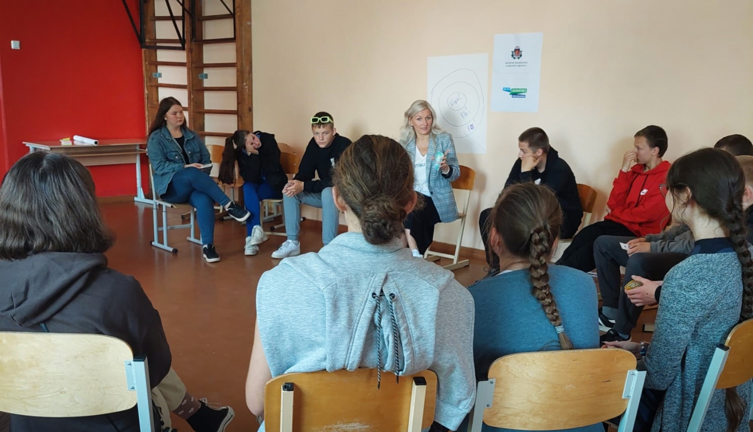 Projekta publicitātes foto - persona runā ar jauniešiem sēžot aplī krēslos