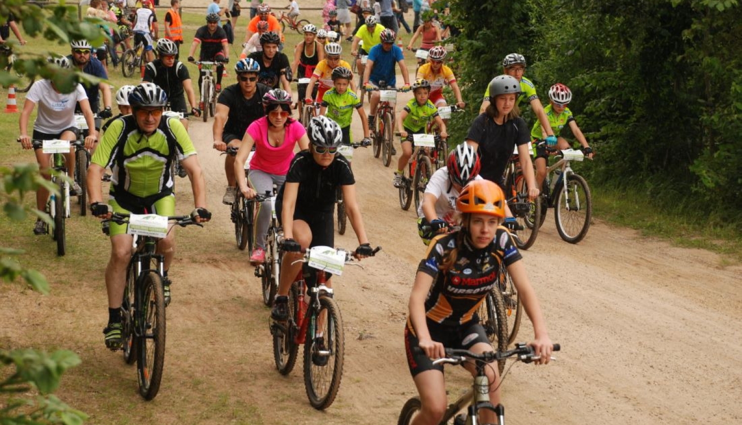 Liels daudzums velosipēdistu dažādos vecumos brauc pa zemes ceļu pretī fotogrāfam