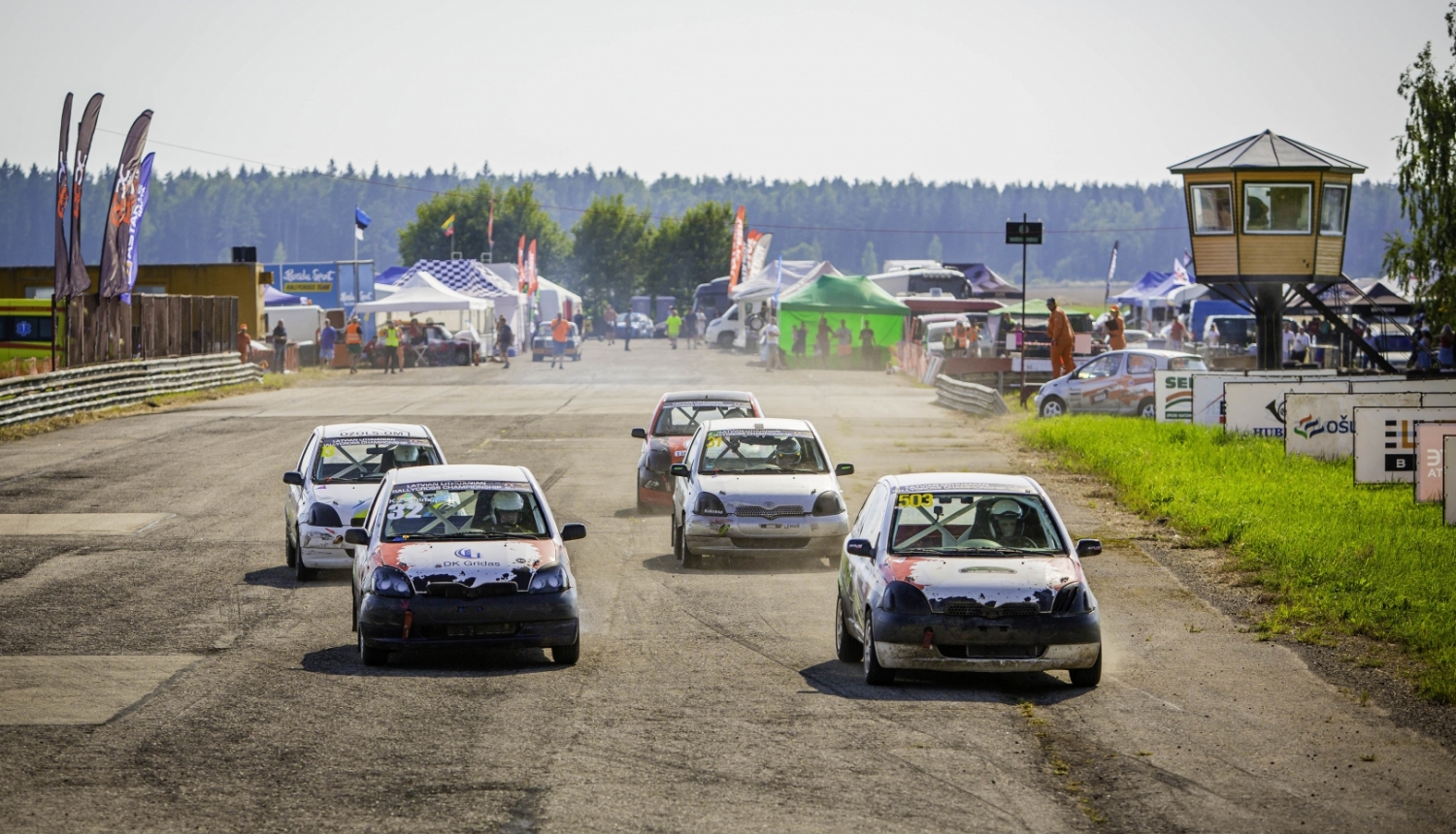 Rallijkrosa automašīnas dodas trasē, fonā tiesnešu būdiņa, teltis un skatītāji